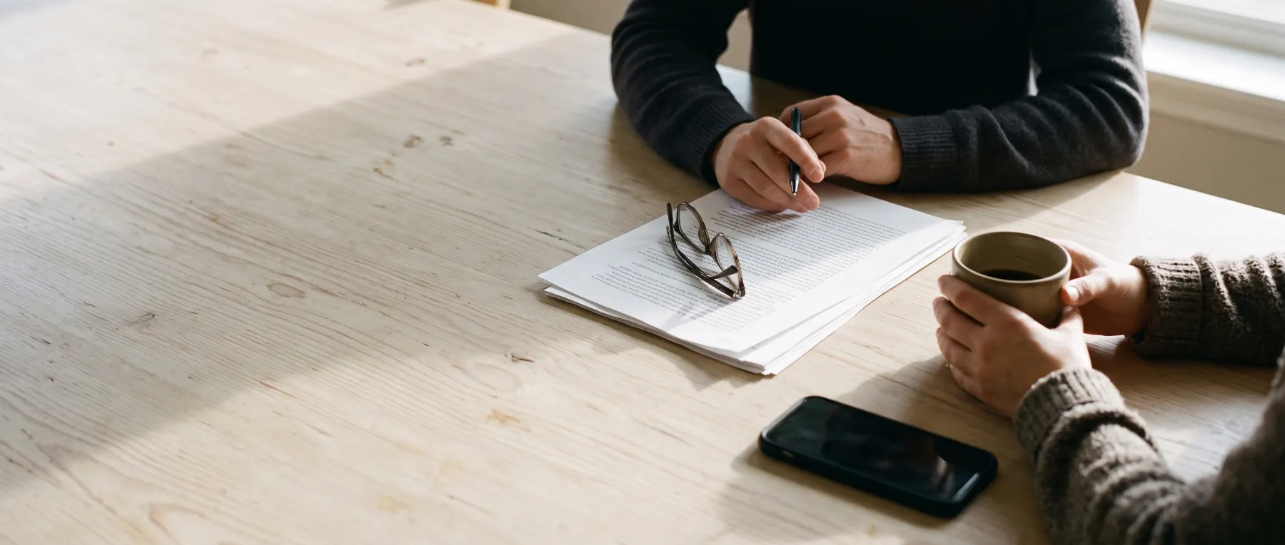 Two people at a pale wooden table, one holding a pen over a printed report, the other holding a coffee mug.